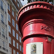 Red postbox with small blue plaque located on the black base