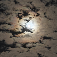 The moon creates a colourful halo through thin clouds, above a field of seedlings, with a road running to the left.