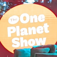 A stage set in readiness for a TV recording. There are two sofas on stage, one of which has a coffee table in front of it with a flask on it. There is a screen on the right explaining it will be live captioned. At the back of the set is the shows logo - the words The One Planet Show in white inside a large red circle. In the foreground we see seats, half of which are filled, with more audience members arriving.