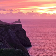 Pink sunset over pink coloured sea with dark green cliffs and a promontory of land stretching out into the sea towards the horizon.