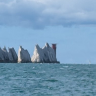 A view from a pebbled beach out across a clear expanse of blue sea to a stack of white cliffs and lighthouse in the far background