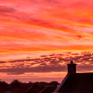 A dramatic morning sky that looks almost as though it is on fire, with dark, shadowy clouds in the distance and a neighbouring house silhouetted against the glowing orange horizon.