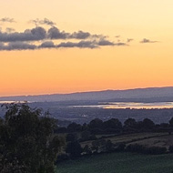 View of green hills, river in the distance, orange tones in the sky as the sunsets and light bounces off the water