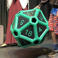A boy smiles on stage while holding a giant inflatable twenty-sided die, with two costumed performers in fantasy dress behind him.